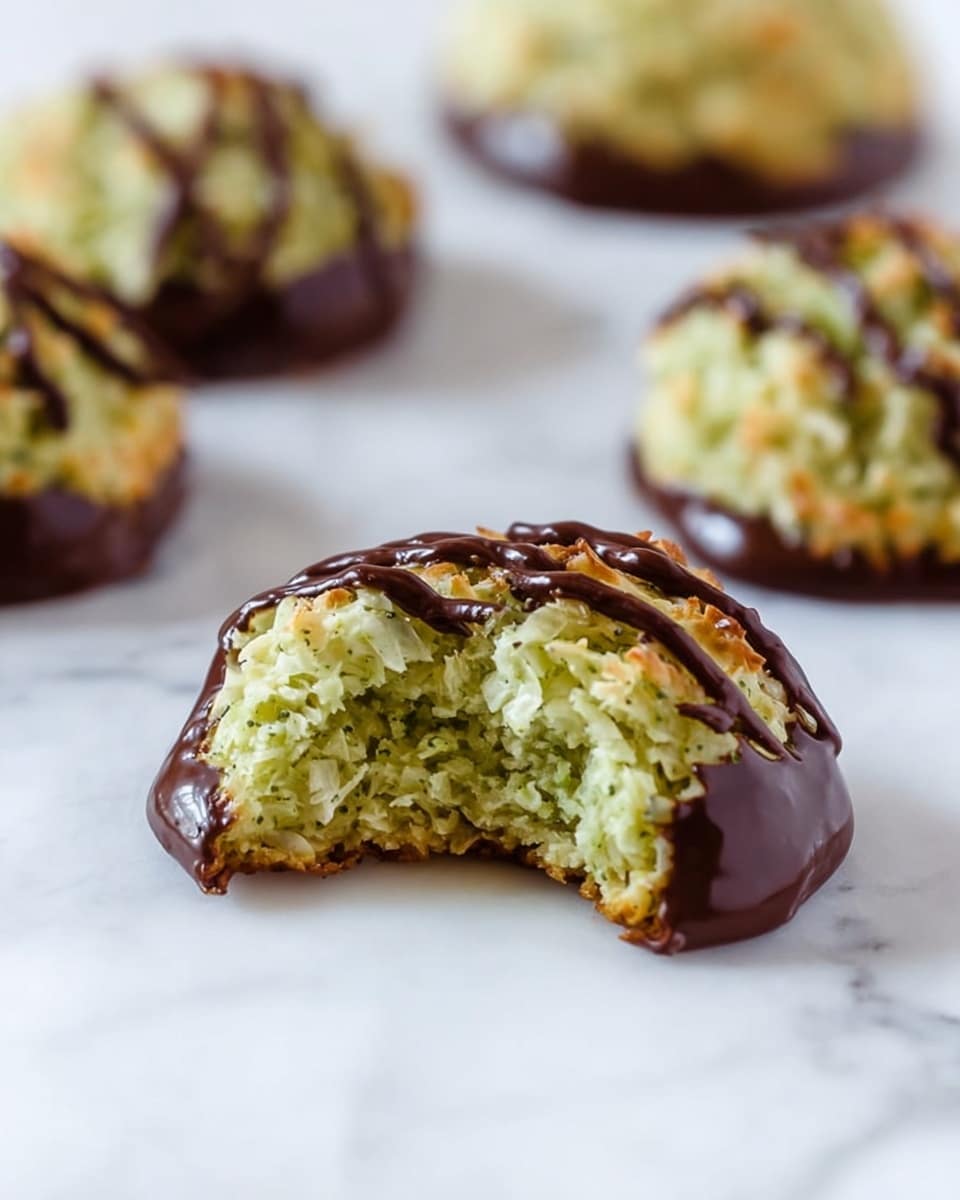 A close-up image of a round cookie with a rough, flaky texture formed from light green coconut flakes, dipped halfway into a shiny dark brown chocolate layer at the bottom. The cookie has a wavy drizzle of the same dark chocolate on top. The front cookie has a large bite taken out, showing the moist, greenish flaky inside. In the background, more cookies of the same kind are softly blurred on a white marbled surface. Photo taken with an iphone --ar 4:5 --v 7