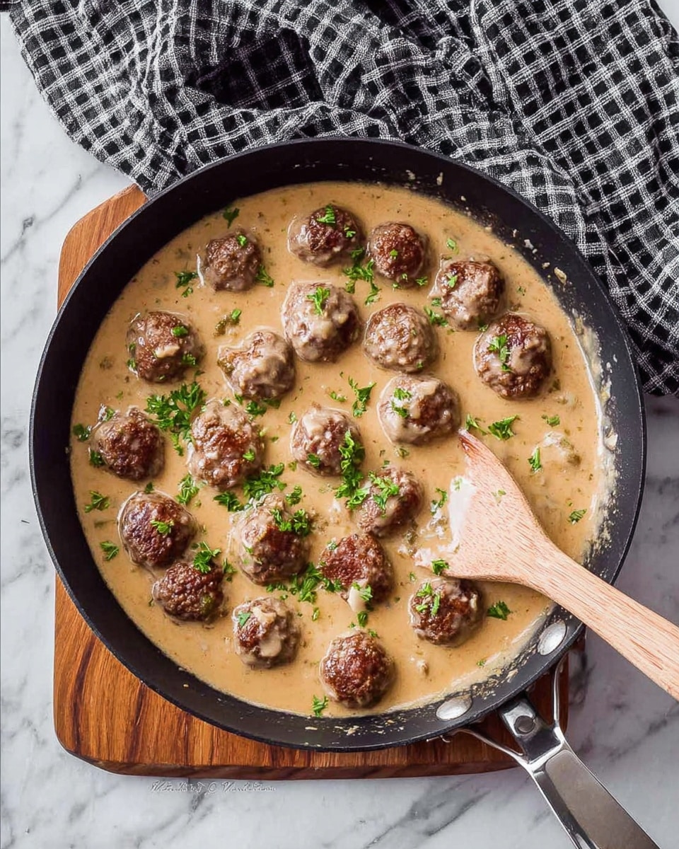 A black skillet filled with two layers: the bottom layer is a creamy light brown sauce with smooth texture, and the top layer is made of round, browned meatballs evenly spread across the sauce, some partially dipped in it. Small green parsley leaves are scattered on top, adding a fresh touch. A wooden spoon is placed on the right side, partly submerged in the sauce, stirring the meatballs. The skillet rests on a white marbled surface with a wooden cutting board underneath part of it and a black and white checkered cloth beside it. Photo taken with an iphone --ar 4:5 --v 7