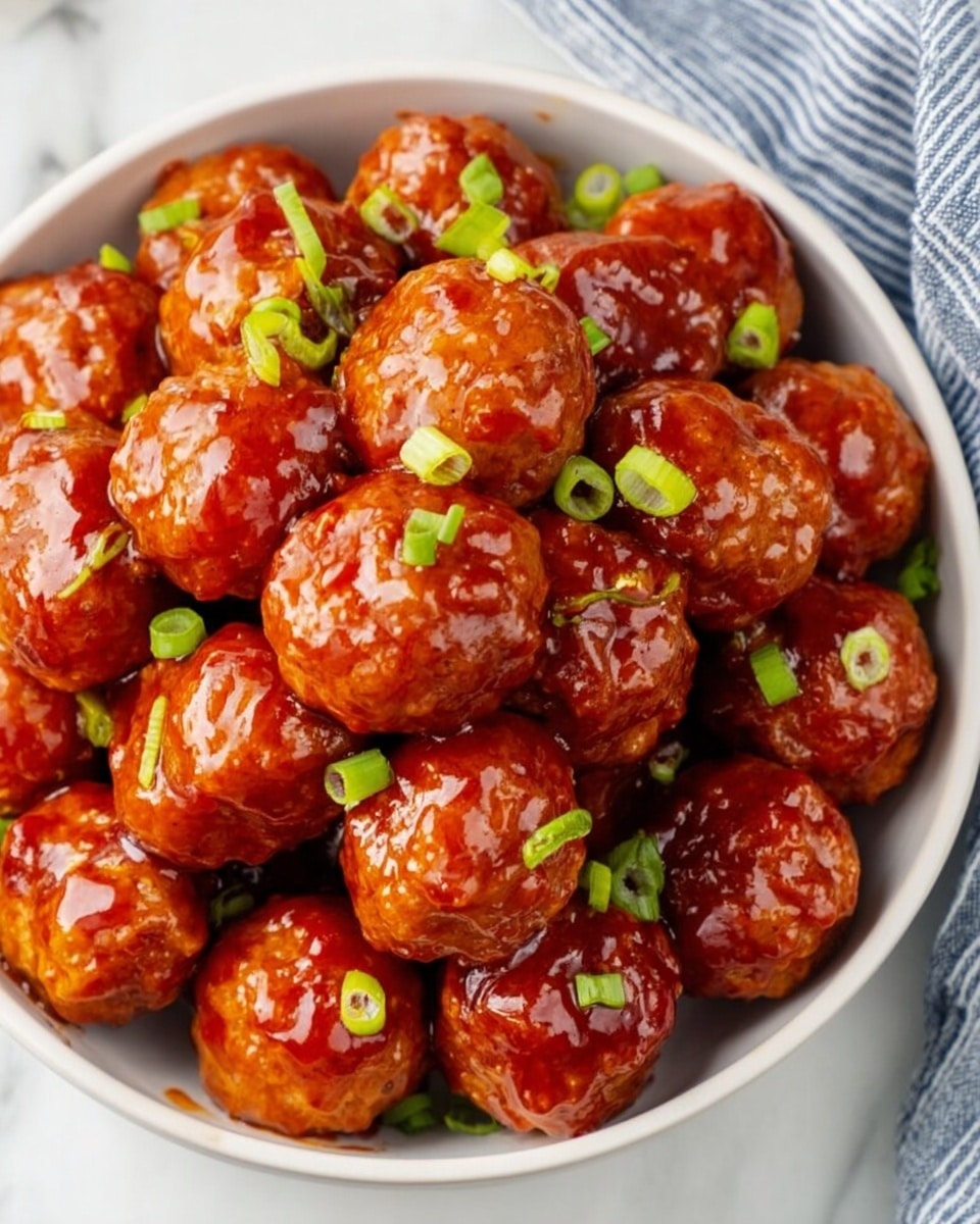 A white bowl filled with shiny, reddish-orange meatballs coated in a thick, glossy sauce, piled high to create about three layers. Bright green small chopped green onion pieces are sprinkled evenly over the top and sides, adding a fresh contrast to the rich sauce. The bowl rests on a white marbled surface with a part of a blue and white striped cloth visible at the top right. The meatballs’ texture is smooth but slightly uneven, showing a cooked and saucy exterior. Photo taken with an iphone --ar 4:5 --v 7