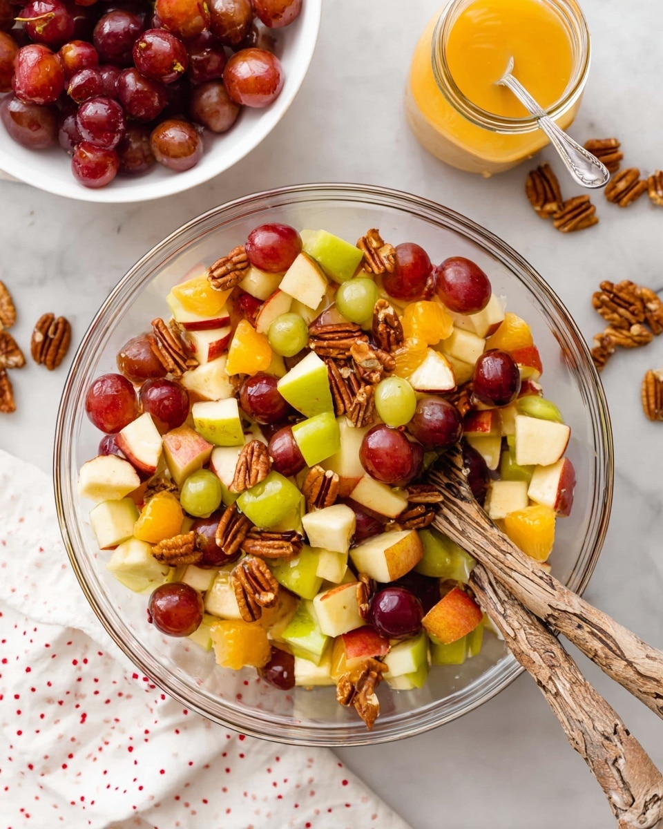 A clear glass bowl filled with a fresh fruit salad sits on a white marbled surface, showing three main layers mixed together: chopped red and green apples with shiny skins, round red grapes that are smooth and firm, and bright orange chunks of oranges. Scattered throughout are textured brown pecan halves and some small dark red dried cranberries. Two long wooden spoons with natural, rough bark-style handles rest inside the bowl, partially buried in the fruit. Nearby, a white bowl filled with more red grapes, loose pecan halves, a glass jar of orange dressing with a spoon, and a white cloth with small red dots are arranged casually. Photo taken with an iphone --ar 4:5 --v 7