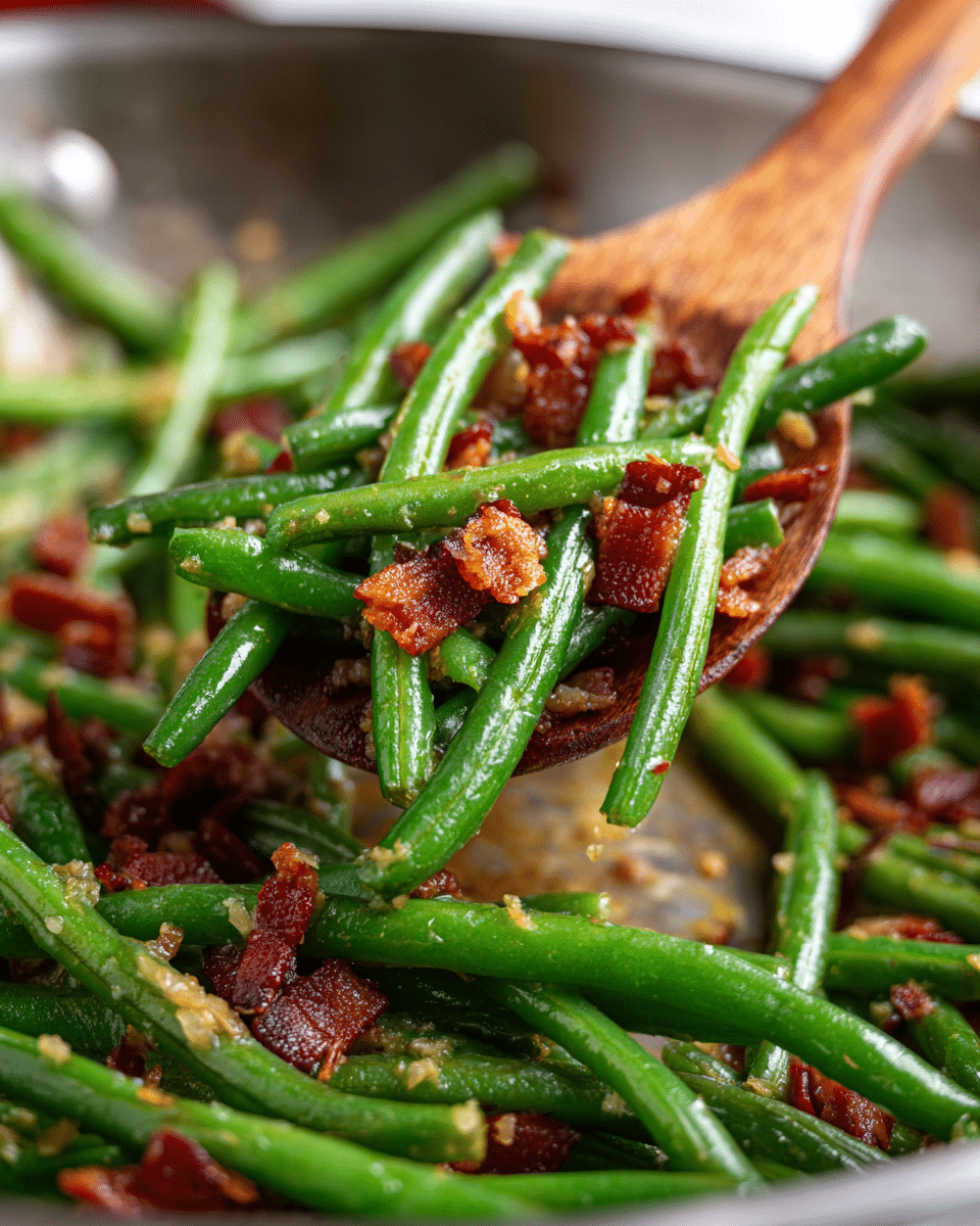 This image shows a close-up of cooked green beans mixed with small pieces of crispy red bacon in a silver pan. A woman's hand is holding a wooden spoon lifting a bunch of shiny green beans coated with a light oily sauce and tiny bits of garlic. The green beans are bright green and smooth, and the bacon pieces are deep red with crispy texture. The pan's surface has some golden-brown cooked oil spots, adding a rich look to the dish. The background is blurred, focusing on the vibrant green beans and bacon. Photo taken with an iphone --ar 4:5 --v 7