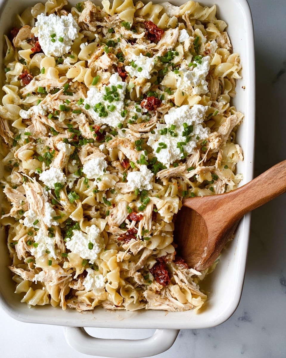 The image shows a close-up of a white ceramic baking dish filled with a pasta dish that has three visible layers. The bottom layer consists of short, curly pasta mixed with shredded chicken that appears light brown and moist. Scattered throughout the pasta are small pieces of sun-dried tomatoes adding hints of red. On top, there is a sprinkling of fresh, finely chopped green herbs, likely chives, adding a pop of color. Finally, dollops of creamy white cheese, possibly ricotta or goat cheese, are spread unevenly across the surface. A wooden spoon rests on the right side of the dish, partially submerged in the pasta. The dish is set on a white marbled surface. Photo taken with an iphone --ar 4:5 --v 7