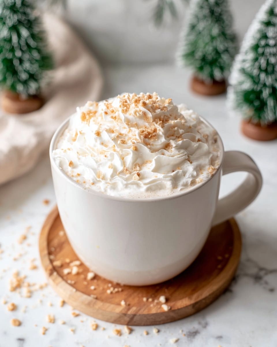 A large white ceramic mug sits on a round wooden coaster, filled to the brim with a creamy beverage topped with a thick layer of whipped cream. The whipped cream is fluffy and white, swirled generously on top, and sprinkled with small, light brown crunchy bits spreading across the cream and slightly falling around the mug. The scene is set on a white marbled surface with soft-focused small green and white decorative trees in the background, giving a cozy, festive feel. Photo taken with an iphone --ar 4:5 --v 7