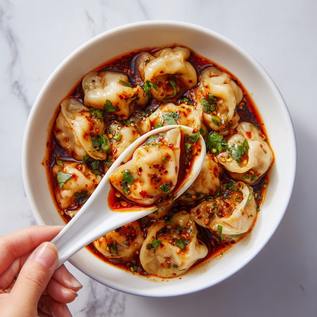 A white bowl filled with many small dumplings covered in a reddish-brown spicy sauce dotted with green herbs. The dumplings are light beige and slightly translucent, showing a soft and smooth texture with some folds. A white spoon held by a woman's hand lifts one dumpling close to the camera, showing the sauce pooling at the bottom of the folds with small pieces of chili and chopped green herbs on top. The background surface is a white marbled texture. photo taken with an iphone --ar 4:5 --v 7