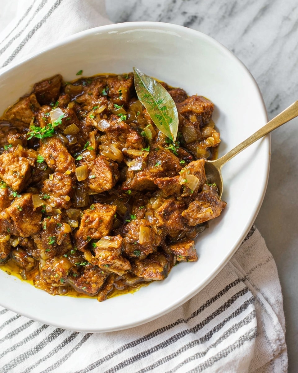 This image shows a white shallow bowl filled with a brown stew made of browned, chunky pieces of meat or tofu, cooked with finely chopped onions and small bits of herbs giving a green color touch. The sauce looks thick and richly coats the pieces, with a visible bay leaf on top. A bronze spoon rests partially inside the bowl, slightly lifting some pieces. The bowl sits on a white marbled surface alongside a white cloth with thin dark stripes. photo taken with an iphone --ar 4:5 --v 7