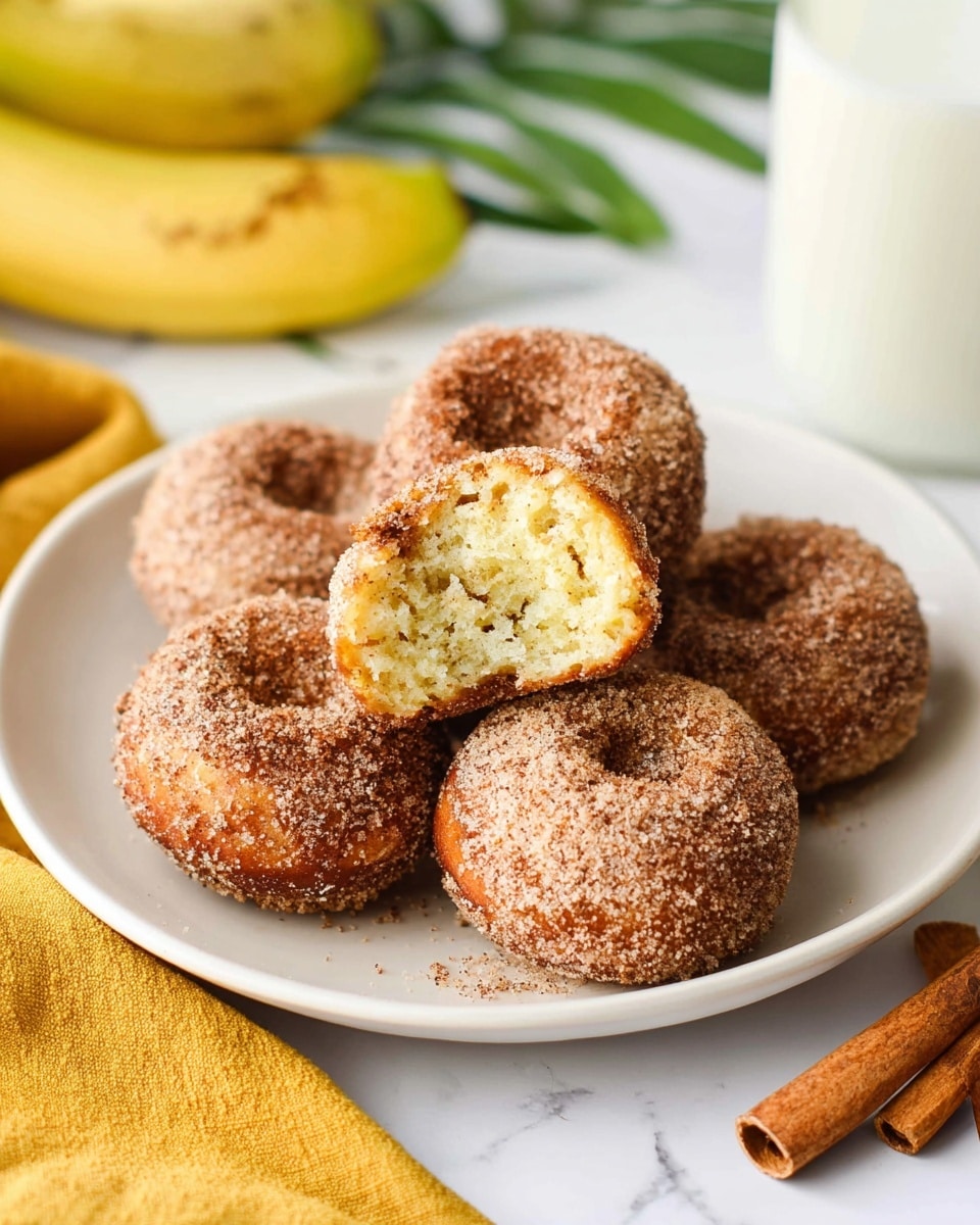 A white plate holds six round cinnamon sugar doughnuts with a rough, grainy outer layer covered in brown sugar and cinnamon. One doughnut is broken in half, showing a soft, fluffy light yellow inside with a textured crumb. The plate sits on a white marbled surface, with a yellow cloth partially visible near the bottom and two cinnamon sticks on the right side. There is a glass of milk slightly blurred in the background, with a banana and some green leaves also softly visible behind the plate. photo taken with an iphone --ar 4:5 --v 7