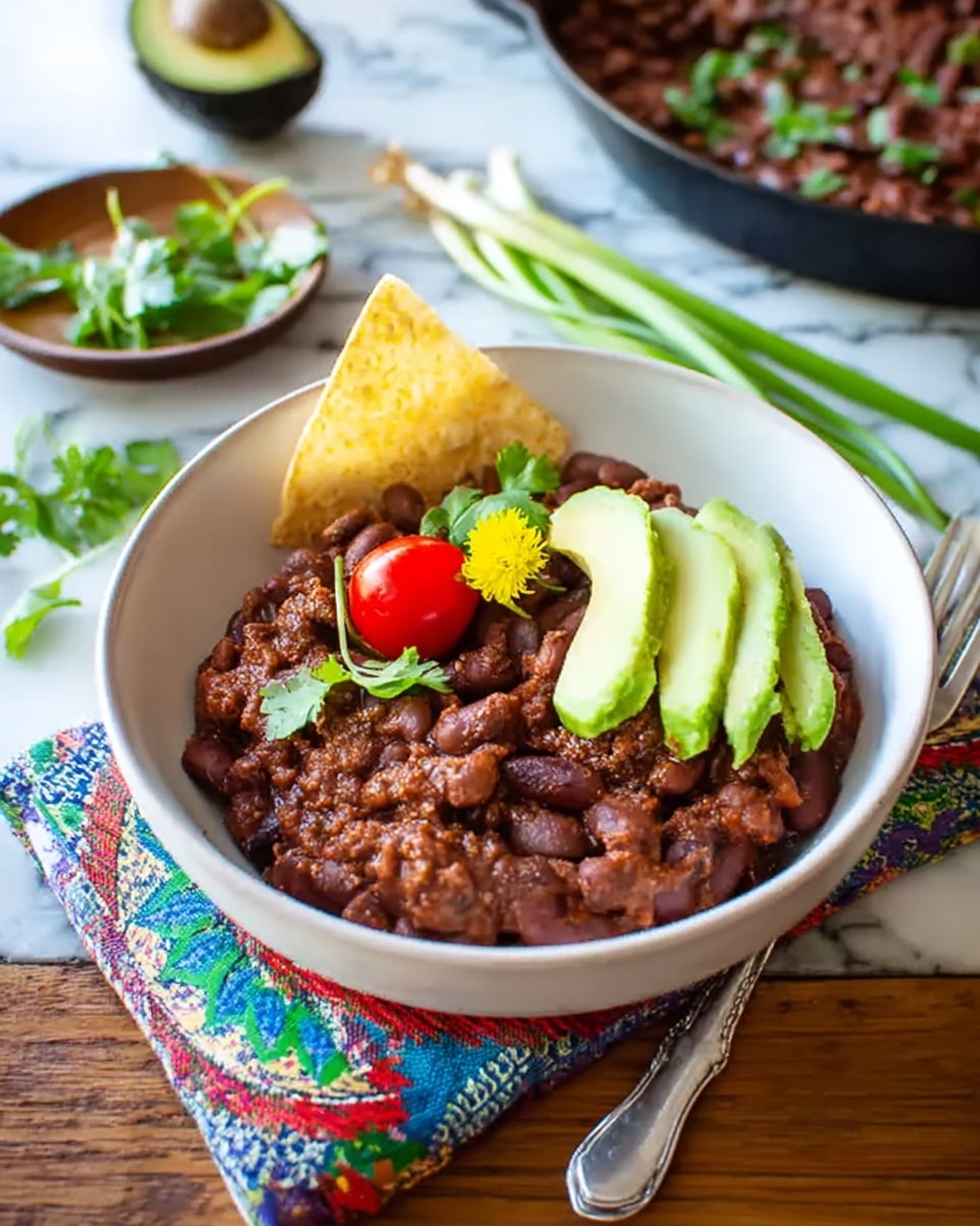 A white bowl filled with cooked red beans that have a soft, textured look, topped with two smooth, green avocado slices placed diagonally. On top of the beans, there is a small bright red cherry tomato, a few green cilantro leaves scattered, and a small yellow flower adding a pop of color. A triangular, pale yellow tortilla chip is tucked into the beans at the back of the bowl. The bowl sits on a colorful patterned cloth over a wooden table, with a silver fork to the right, and an avocado half and some green onions nearby. In the background, there is a pan filled with more beans garnished with green herbs. The whole scene is set on a white marbled surface. photo taken with an iphone --ar 4:5 --v 7