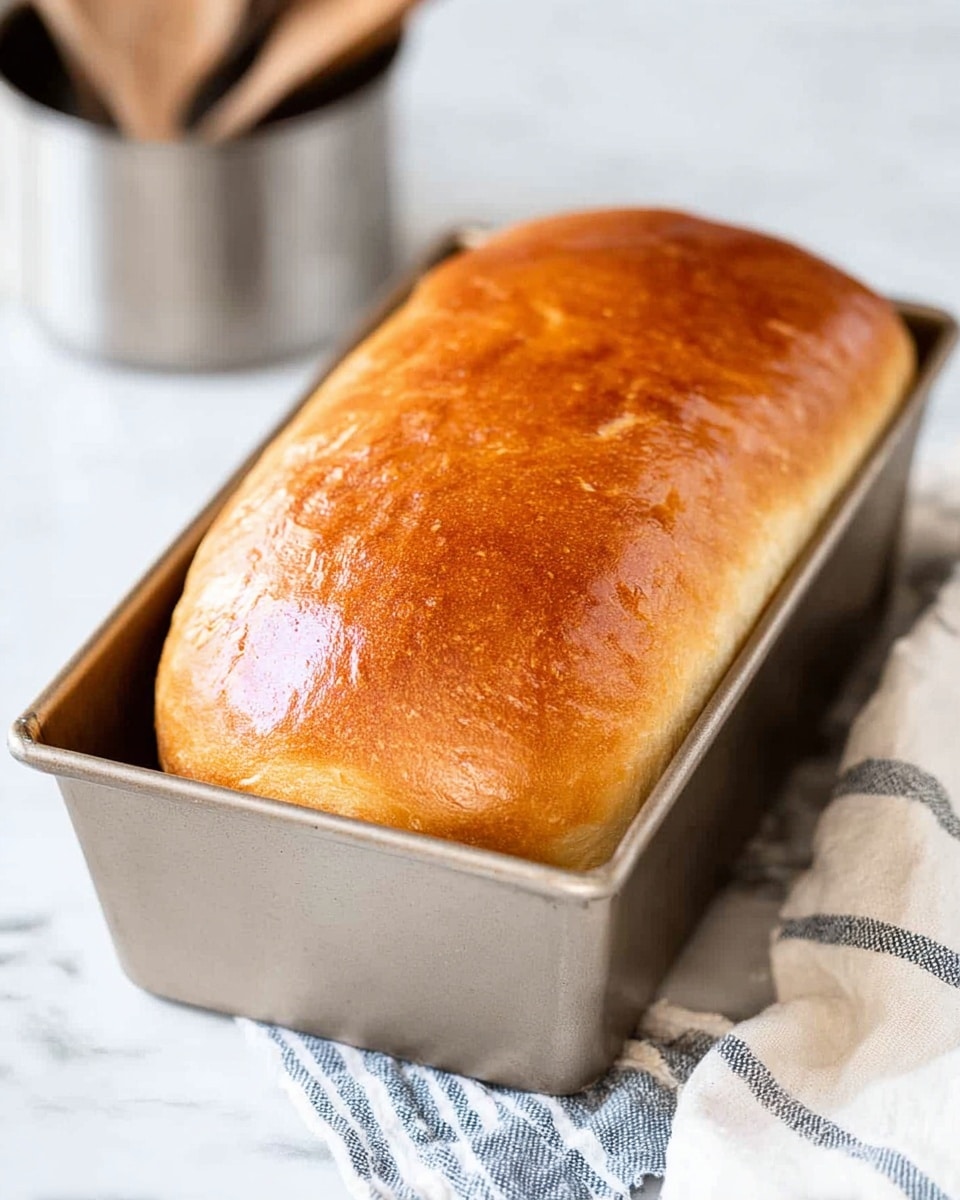 The image shows a golden brown loaf of bread freshly baked inside a silver metal baking pan. The loaf has a smooth, shiny top crust that looks soft and slightly glossy with a few small cracks. The bread fills the pan well and rises just above the edges. The background is a white marbled texture with a blurred metallic cup and wooden spoons visible in the distance. A white and blue striped cloth is partially seen beside the pan. photo taken with an iphone --ar 4:5 --v 7