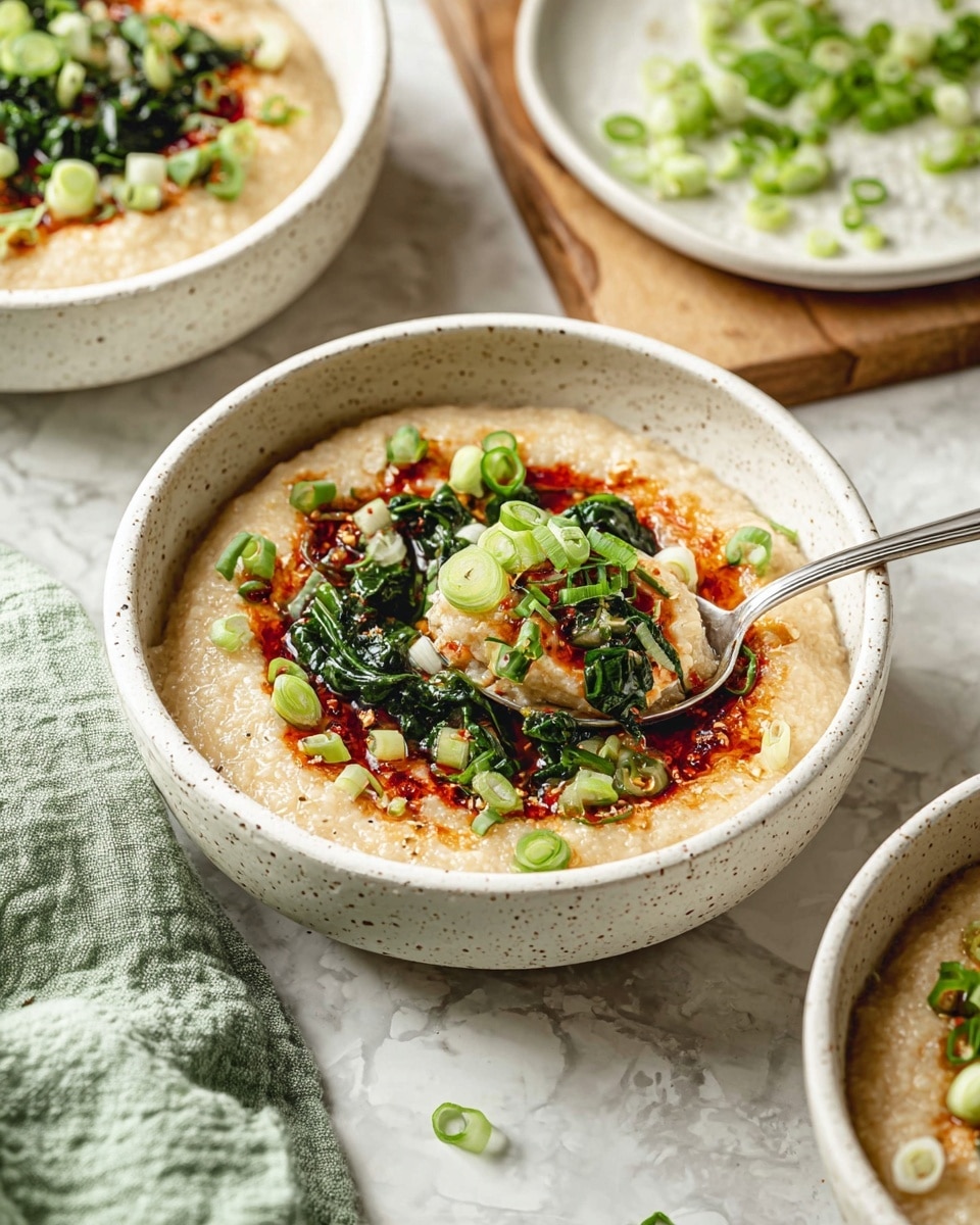 A close-up view of a creamy, beige porridge served in a white speckled bowl. The porridge is spread evenly across the bowl as the base layer, topped with a glossy dark green leafy vegetable mix in the center, layered with thin light green scallion rings scattered on top. There is a vibrant red chili oil drizzle around the greens, creating bright streaks on the porridge. A silver spoon rests inside the bowl, partially scooping the porridge. In the background, two similar bowls with the same dish are faintly visible, placed on a white marbled surface, one on a wooden board. A light green cloth is seen in the lower left corner. Photo taken with an iphone --ar 4:5 --v 7