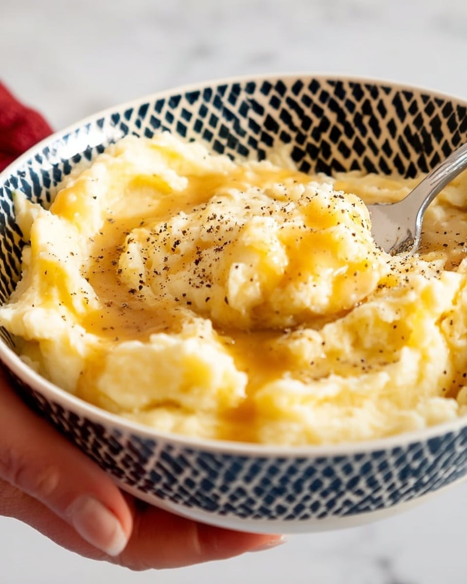 A close-up of a bowl filled with creamy mashed potatoes. The mashed potatoes are light yellow with a smooth yet slightly chunky texture, topped with a drizzle of golden brown gravy and sprinkled with black pepper. The bowl is white with a black geometric pattern on the outside. A woman's hand is holding the bowl on the left side, and a spoon is partially visible on the right side inside the bowl. The background features a white marbled texture. photo taken with an iphone --ar 4:5 --v 7