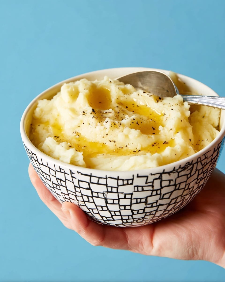 A close-up shows a woman’s hand holding a white bowl with a black square pattern filled with creamy mashed potatoes. The mashed potatoes are light yellow with a smooth, slightly fluffy texture, topped with melted butter that creates shiny, golden streaks. Small specks of black pepper are sprinkled across the top, adding contrast. A silver spoon rests inside the bowl on the right side. The background is a plain blue color. photo taken with an iphone --ar 4:5 --v 7