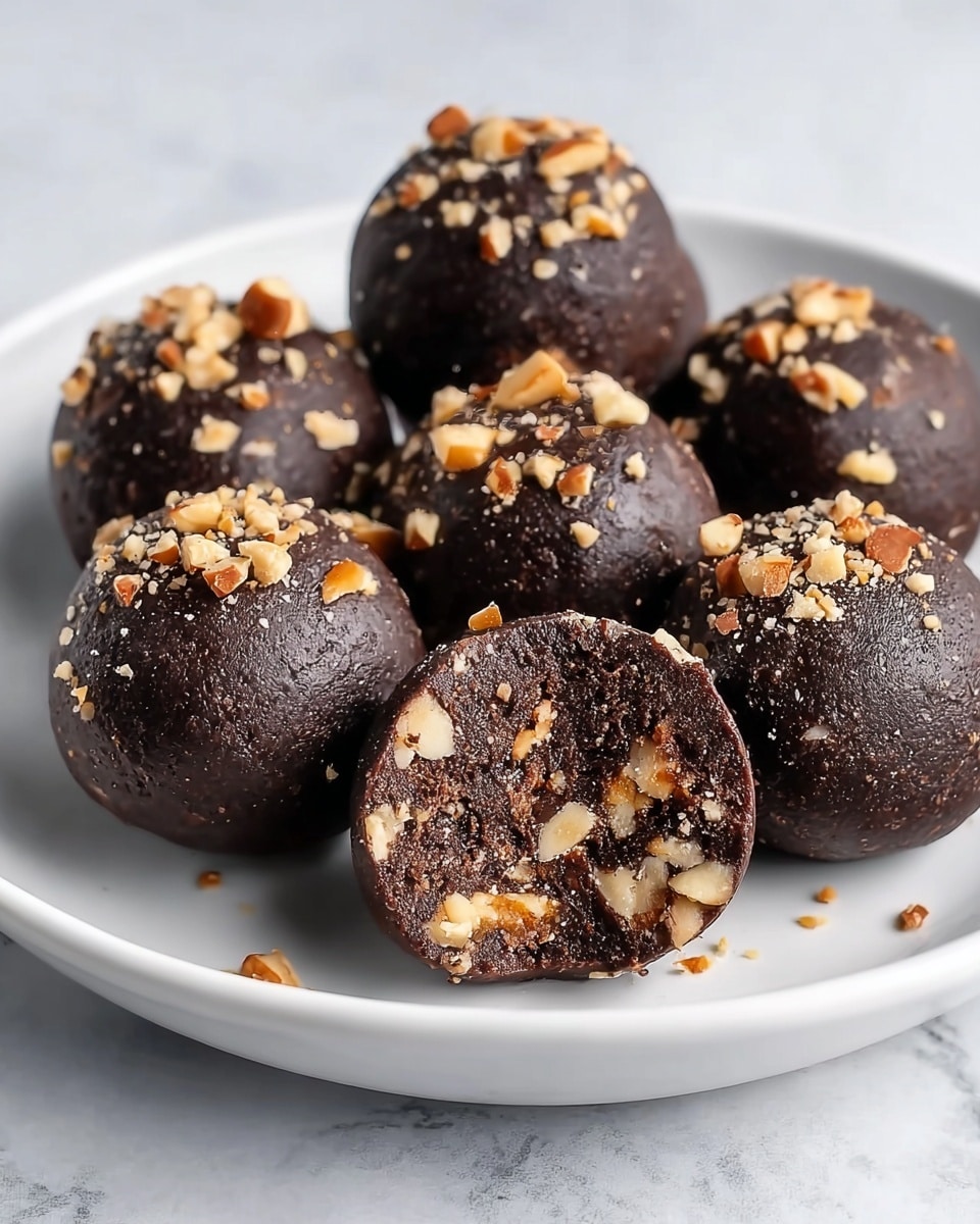 The image shows a white plate with seven dark brown chocolate balls, each sprinkled with small pieces of light brown nuts on their surfaces. One chocolate ball is cut in half and placed at the front, revealing a dense, textured inside filled with bits of nuts that are light brown and embedded throughout the dark chocolate mixture. The plate is set on a white marbled surface. photo taken with an iphone --ar 4:5 --v 7