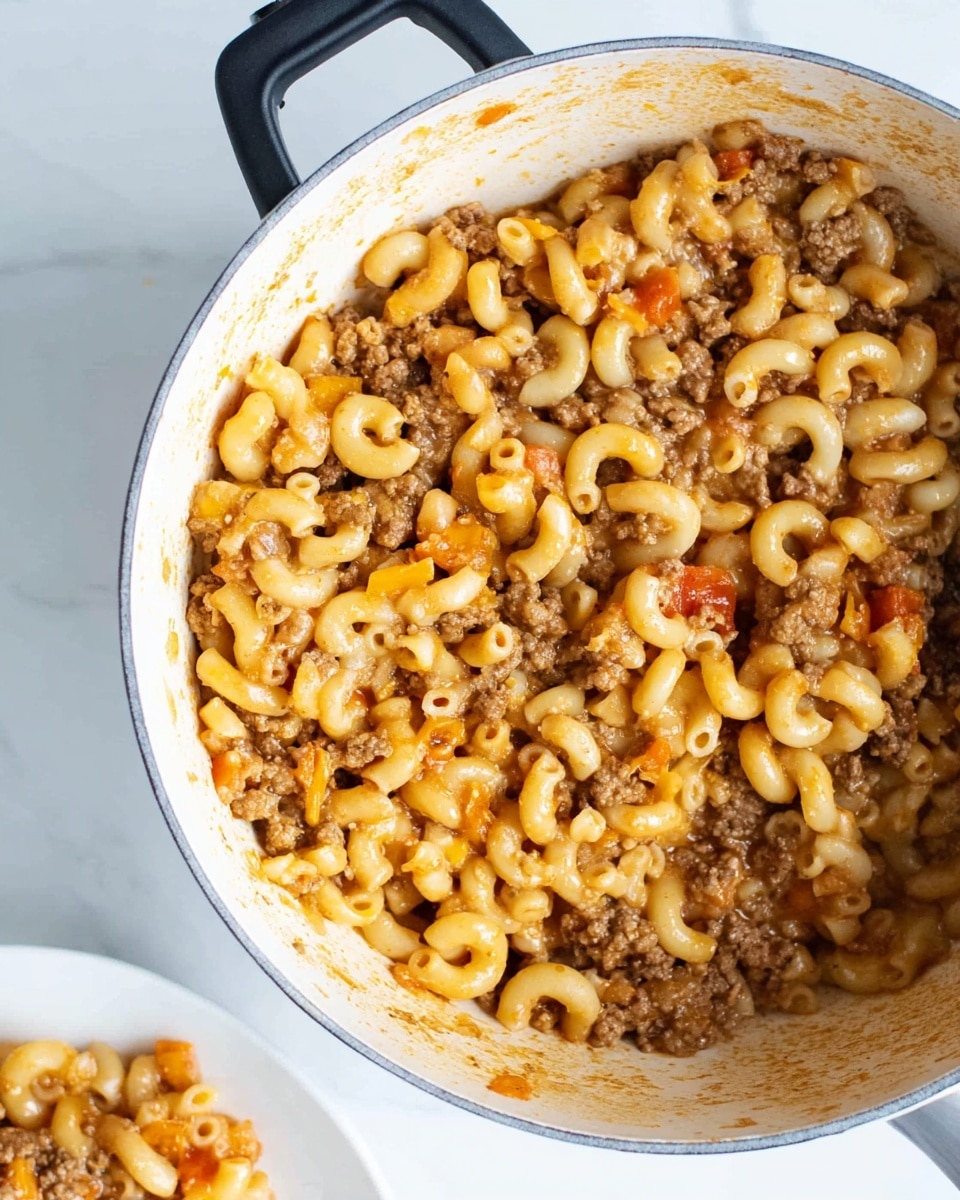 A close-up view of a white pot filled with cooked elbow macaroni pasta mixed with browned ground beef and small pieces of orange cheddar cheese, all coated in a light brown sauce with small bits of tomato visible. The pot shows some sauce residue on the sides, and a portion of the macaroni beef mix is partially served on a white plate visible at the bottom left. The pot has a black handle on top and sits on a white marbled surface. Photo taken with an iphone --ar 4:5 --v 7