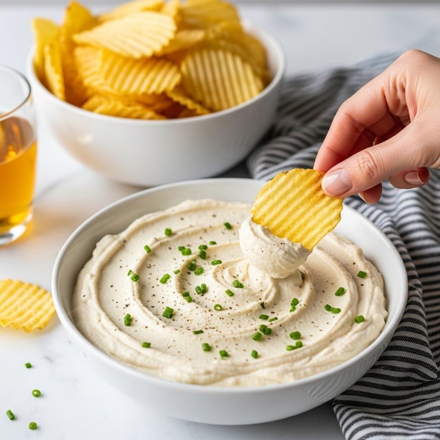 A white bowl filled with a creamy, thick, off-white dip spread smoothly with soft swirls across the surface, topped with small green chopped chives scattered evenly and a light sprinkle of black pepper. A woman's hand is dipping a golden, ridged potato chip into the dip, creating a small dip mark. Behind the bowl, there is a white bowl filled with more golden potato chips with a ridged texture. The setting includes a white marbled textured surface, with a black and white striped cloth placed near the bowl and a glass of light brown liquid in the background. Photo taken with an iphone --ar 4:5 --v 7