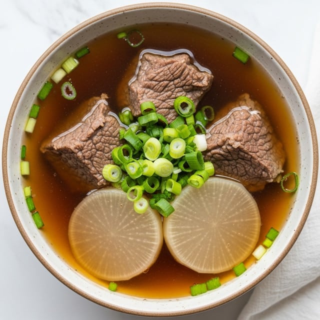 A close-up view of a bowl filled with clear brown broth containing three pieces of tender brown beef with visible texture and two large white radish slices nestled inside. Bright green chopped scallions are sprinkled on top of the beef and radish, adding a fresh pop of color. The bowl is white with a speckled rim, resting on a white marbled surface with part of a white cloth visible nearby. The light shines softly, highlighting the textures and colors of the soup. photo taken with an iphone --ar 4:5 --v 7