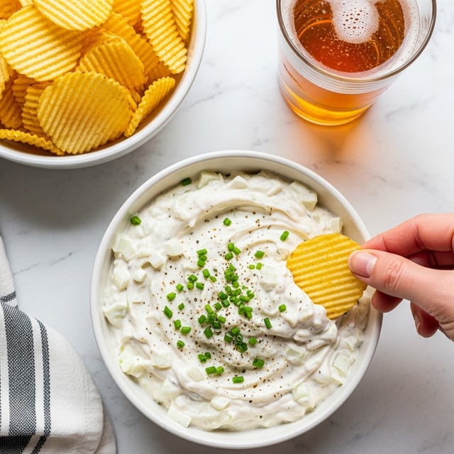 A white bowl filled with creamy white onion dip mixed with visible chunks of onion, topped with chopped green chives and a sprinkle of black pepper, with a woman's hand holding a ridged potato chip dipping into the bowl on the right side. Above and to the left, another white bowl is filled with more ridged potato chips in a golden-yellow color. On the upper right corner, a glass of amber-colored beer with bubbles is visible. The whole scene is set on a white marbled surface with part of a black and white striped cloth on the lower left side. Photo taken with an iphone --ar 4:5 --v 7