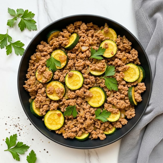 The image shows a black round pan filled with a cooked mixture made of light brown ground meat and sliced green zucchini pieces. The dish is garnished with bright green parsley leaves scattered on top. The meat has a juicy texture with some visible small chunks of onions mixed in. The colors contrast well with the white marbled surface beneath the pan and scattered parsley around it. There is a hint of seasoning visible in small black and white grains near the lower left. The scene includes a gray cloth on the right side, adding softness to the setting. photo taken with an iphone --ar 4:5 --v 7