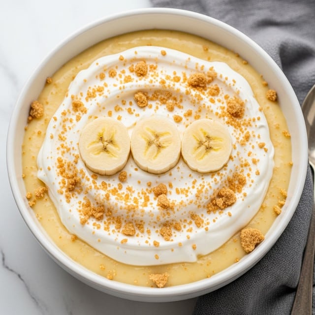A white bowl filled with a creamy, light yellow pudding as the base layer, topped with a smooth layer of white whipped cream slightly swirled on top. Three banana slices with a soft yellow color and visible seeds are placed in a row in the center. Crushed light brown crumbs are generously sprinkled over the whipped cream and around the bananas. The bowl sits on a white marbled surface and a gray cloth is visible nearby. photo taken with an iphone --ar 4:5 --v 7