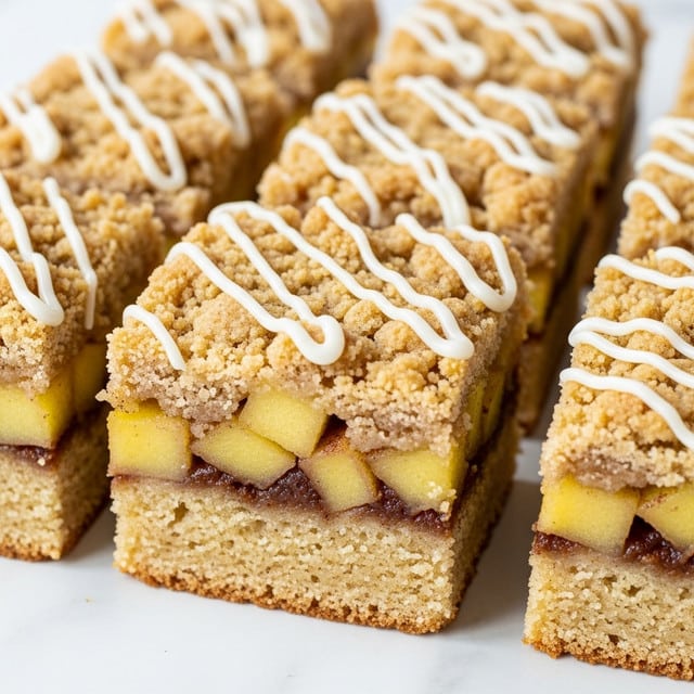 The image shows several square pieces of crumb cake with a crumbly, golden-brown top layer sprinkled with bits of cinnamon and apple chunks. Each piece has two main layers: a thick, soft, light yellow cake base that has a few swirls of cinnamon and apple chunks embedded inside, and a crumbly, textured top layer with a drizzle of white icing over it, creating a contrast with the cake. The squares are arranged close together on a dark surface. photo taken with an iphone --ar 4:5 --v 7