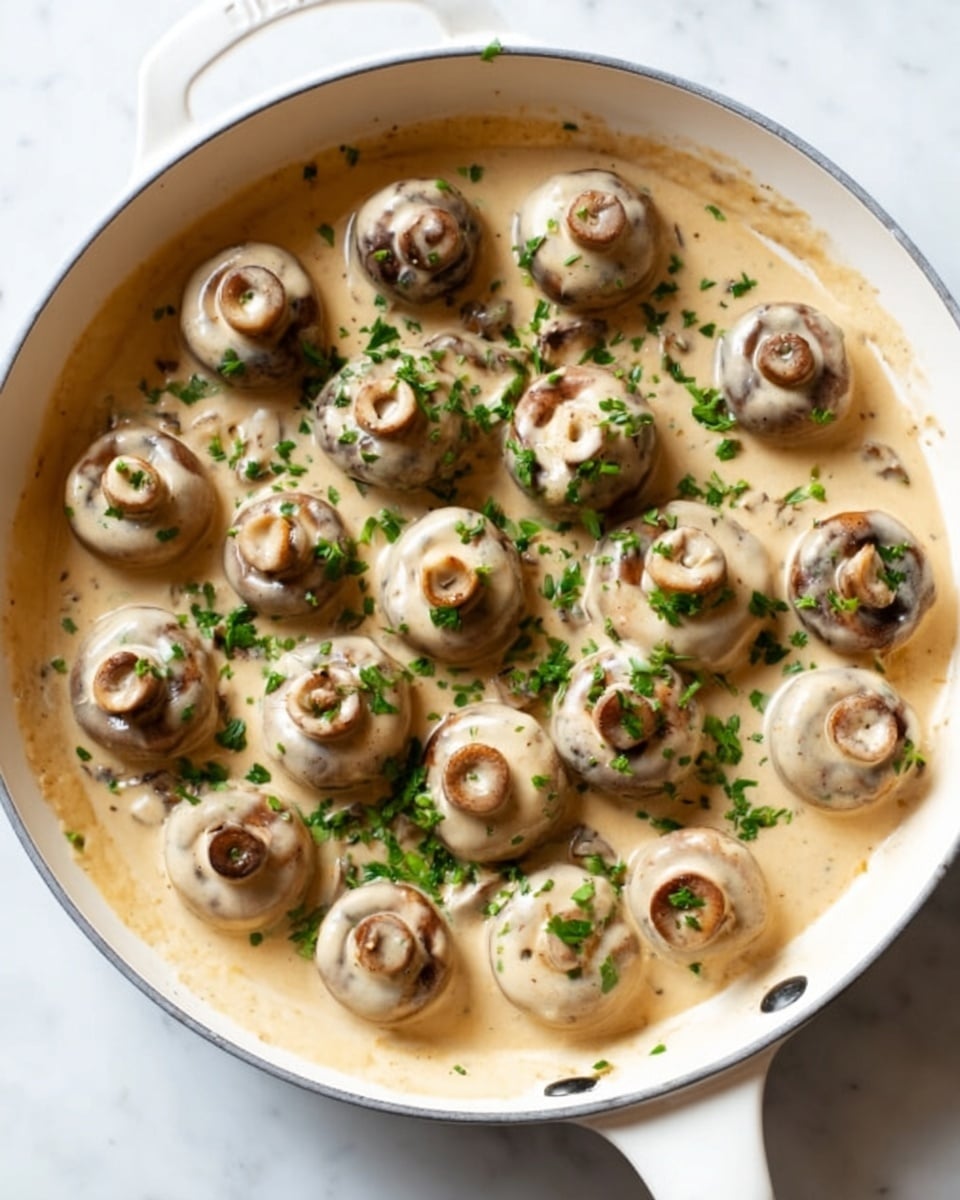 A black cooking pan filled with whole white mushrooms cooking in a creamy light brown sauce, with small green herb pieces sprinkled on top and some black pepper visible. The mushrooms are arranged evenly across the pan, all covered partly by the sauce that has a smooth texture. The pan is on a white marbled surface. Photo taken with an iphone --ar 4:5 --v 7