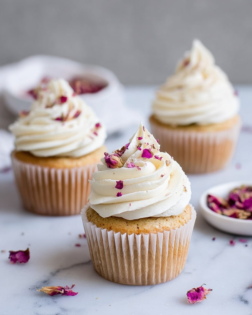 Three vanilla cupcakes with light brown cake bases wrapped in beige paper liners are arranged on a white marbled surface. Each cupcake has a tall swirl of smooth, bright white frosting on top, decorated with small dried rose petals in shades of pink, purple, and red, scattered gently over the frosting and surface. A white bowl filled with more dried petals is blurred softly in the background. photo taken with an iphone --ar 4:5 --v 7