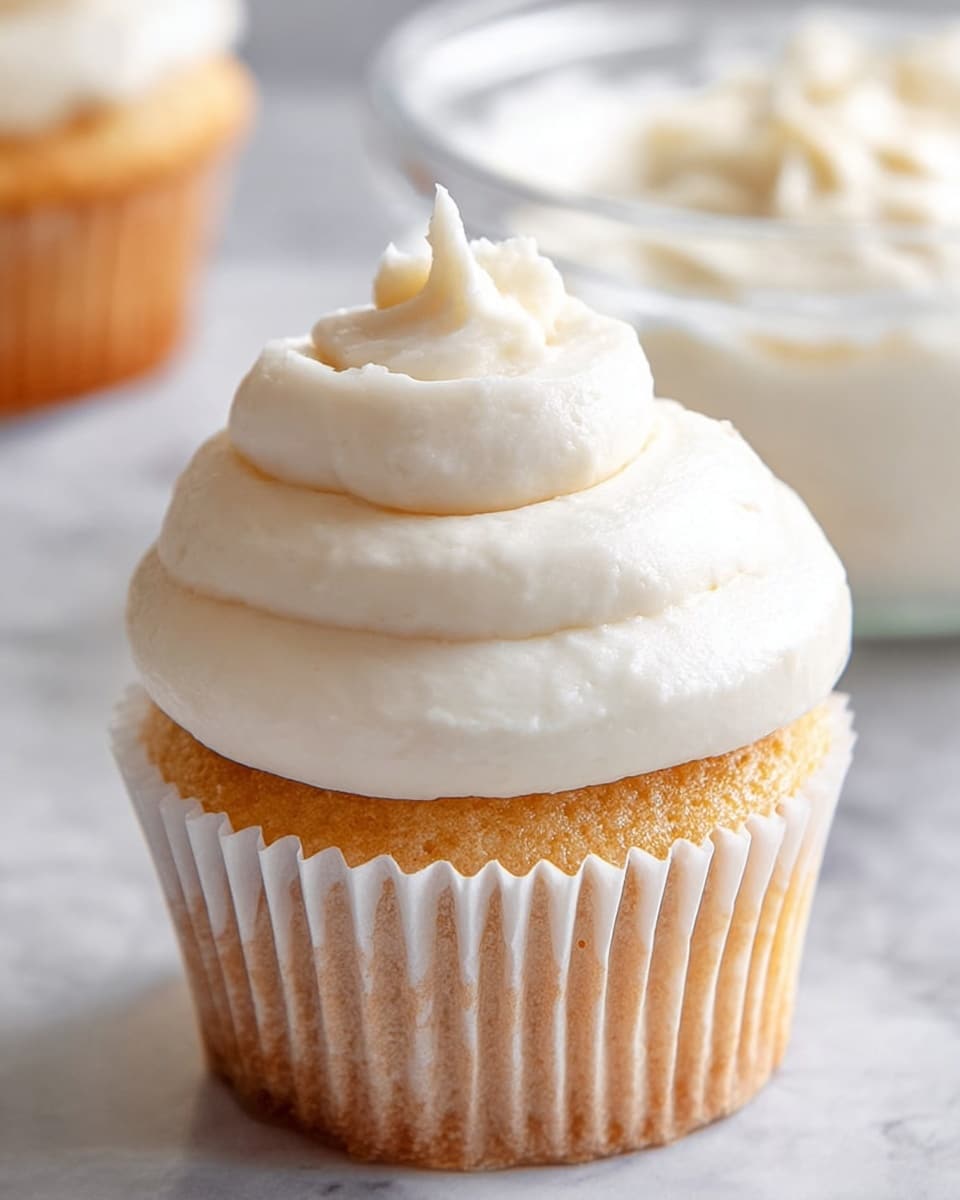 A cupcake with a light golden-brown base wrapped in a white paper liner is topped with three thick, smooth layers of creamy white frosting, each layer slightly larger than the one above it, forming a soft spiral with a pointed peak at the top. The cupcake is placed on a white marbled surface with a glass bowl of the same creamy white frosting blurred in the background. The overall look is soft and inviting, with the frosting showing a slightly glossy texture. photo taken with an iphone --ar 4:5 --v 7