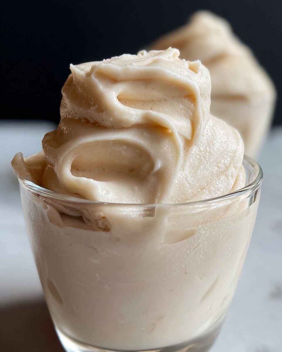 A clear glass cup filled to the top with a thick, creamy, pale beige paste that is smooth and slightly shiny. The paste forms soft peaks and swirls above the rim of the cup, giving it a rich and dense appearance. In the background, there is another similar container with the same creamy substance, set against a dark and simple backdrop with a white marbled surface below. photo taken with an iphone --ar 4:5 --v 7