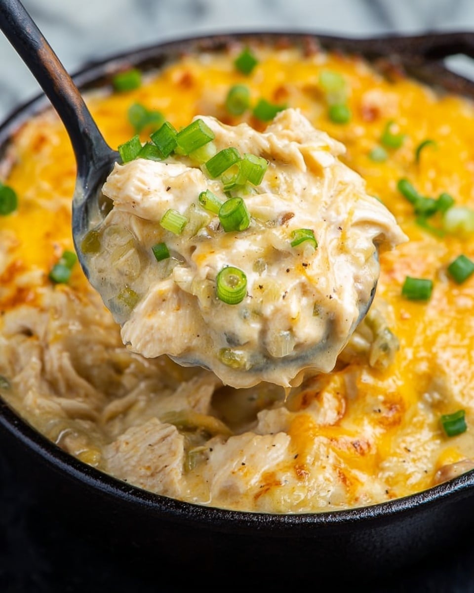 A close-up of a creamy casserole being scooped by a black spoon from a cast iron skillet on a white marbled surface. The dish has layers of shredded white chicken mixed with a smooth, thick, light beige sauce, topped with melted golden-yellow cheese that looks gooey and slightly browned. Small bright green onion pieces are sprinkled on top, adding a fresh color contrast. The texture appears rich and thick, with the cheese stretching slightly as the spoon lifts a portion. Photo taken with an iphone --ar 4:5 --v 7