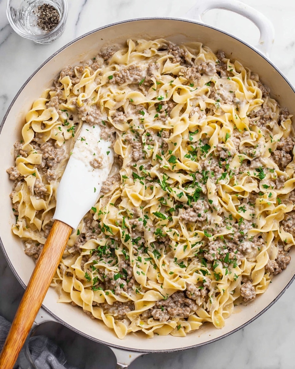 A white pan filled with a creamy pasta dish made of three main layers: light beige cooked egg noodles with a slightly curly texture, browned ground meat mixed evenly throughout, and a smooth, white creamy sauce coating the noodles and meat. Small bits of chopped green herbs are sprinkled on top for color contrast. A white spatula with a wooden handle is placed inside the pan, lifting some of the pasta mixture. The pan sits on a white marbled surface with a folded green and white patterned cloth nearby and a salt shaker in the upper right corner. Photo taken with an iphone --ar 4:5 --v 7