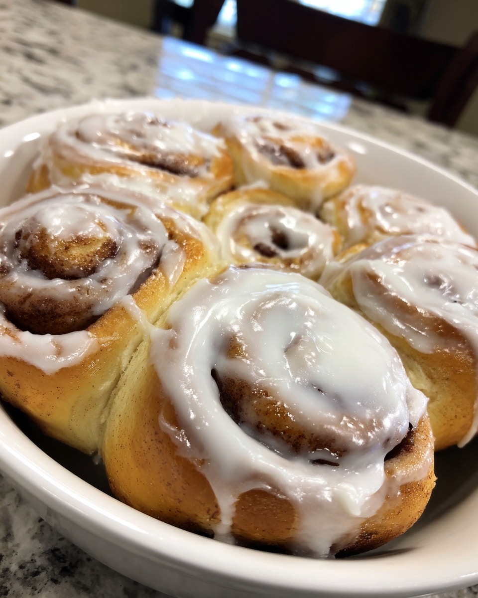 The image shows close-up views of soft cinnamon rolls covered in smooth white cream cheese frosting. Each roll has a spiral pattern with a golden-brown dough layer beneath, visible around the edges and through parts of the frosting. The frosting looks thick and creamy, spread evenly on top of each roll, with some texture on the surface. The rolls are placed closely together in a white dish set on a white marbled surface. photo taken with an iphone --ar 4:5 --v 7