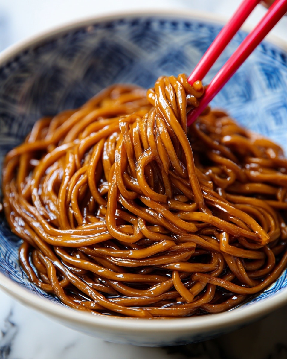 The image shows a close-up of a bowl of glossy, dark brown noodles coated in a thick sauce. The noodles are densely packed and slightly twisted, sitting inside a white bowl with a blue speckled pattern on the inside. A pair of red chopsticks is lifting a clump of noodles from the bowl, creating depth and focus on the shiny texture of the sauce clinging to the strands. The background is a white marbled texture, giving a clean and light contrast to the rich colors of the noodles and bowl. photo taken with an iphone --ar 4:5 --v 7