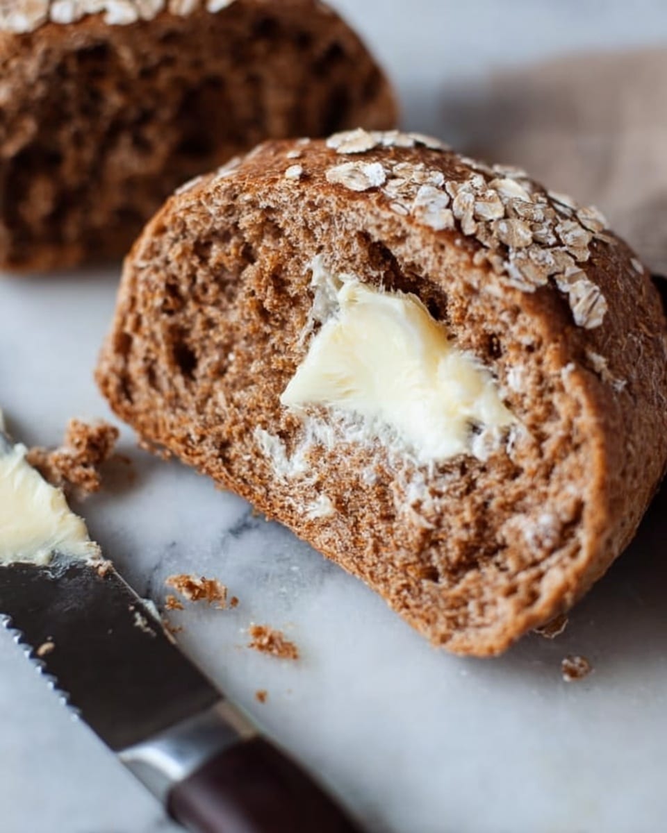 A close-up image of a broken whole wheat bread roll on a white marbled surface, showing one half with a thick layer of melting butter spread unevenly on the rough, brown, dense interior crumb, with the other half of the roll empty beside it. The exterior crust is medium brown and sprinkled with white oats. A serrated knife lies next to the bread, and small crumbs are scattered around. Photo taken with an iphone --ar 4:5 --v 7
