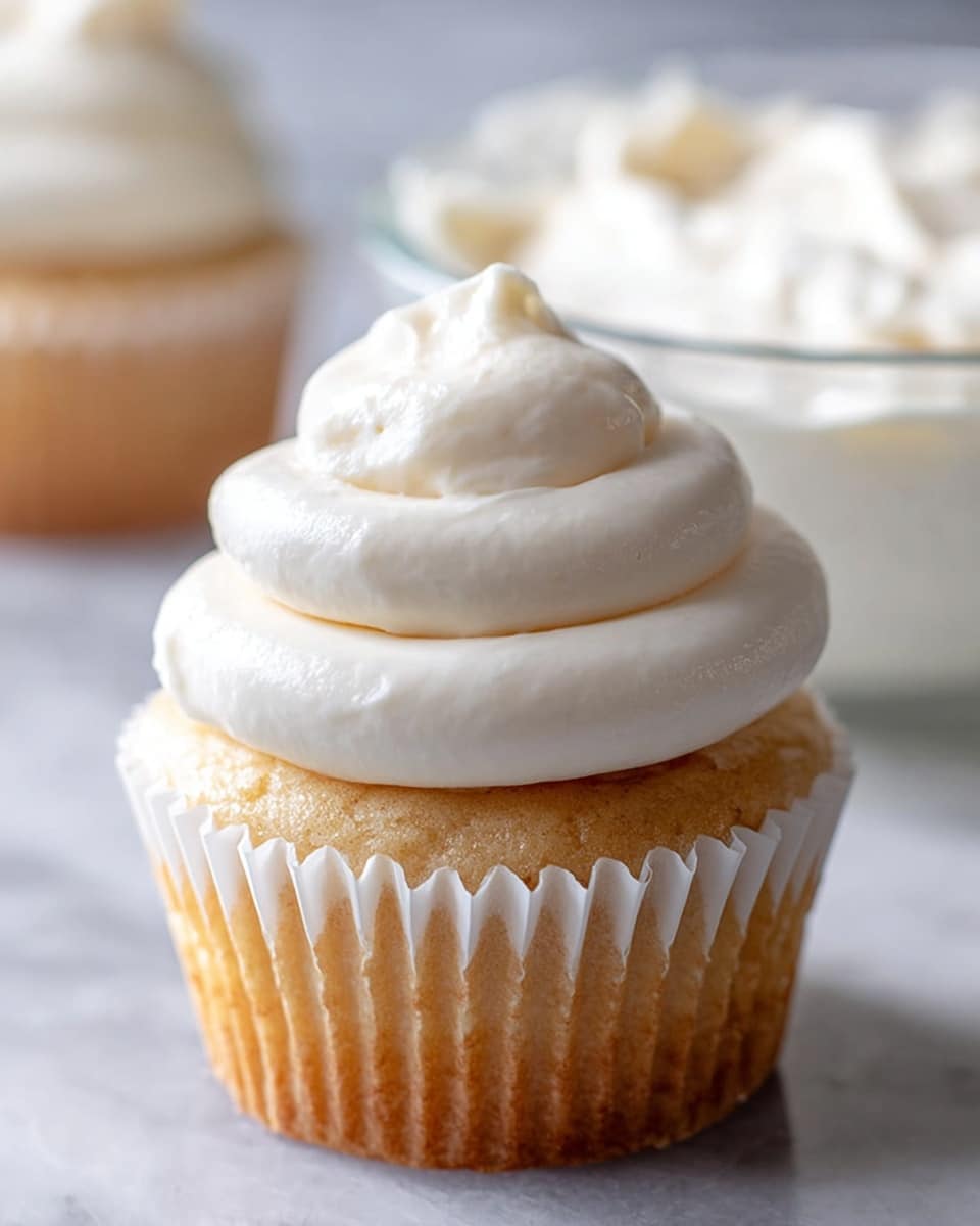 A close-up image of a vanilla cupcake with three thick layers of smooth, creamy white frosting swirled on top, forming a soft peak at the center; the cupcake is in a white ridged paper liner with a golden-brown base visible beneath the frosting, all placed on a white marbled texture surface with a clear glass bowl of extra frosting slightly blurred in the background; photo taken with an iphone --ar 4:5 --v 7