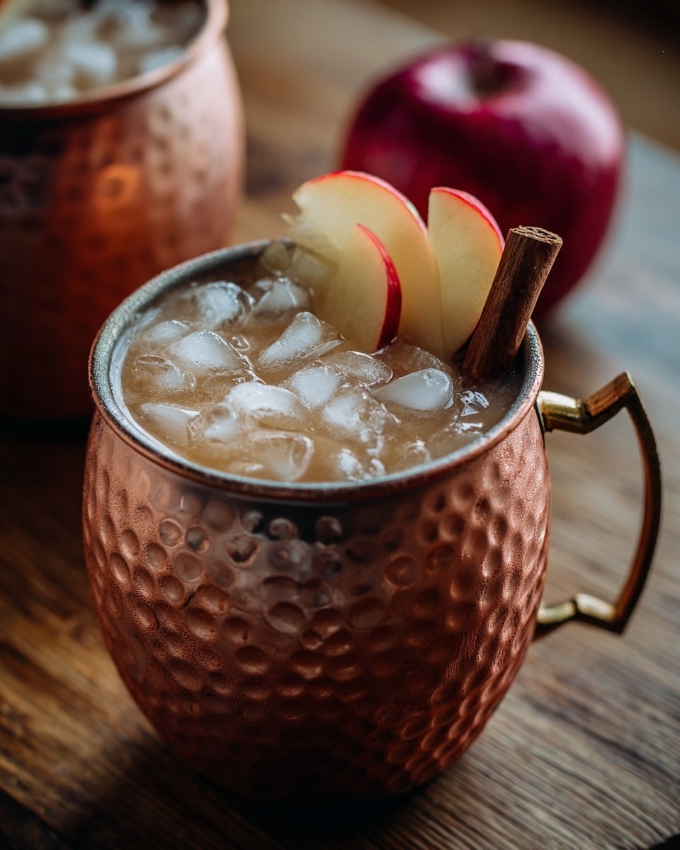 A hammered copper mug filled with a light brown iced drink topped with many clear ice cubes, two apple slices with red edges placed on one side inside the mug, and a dark brown cinnamon stick standing upright beside the apple slices. The mug sits on a wooden surface with a blurred whole red apple and a second copper mug in the background. Photo taken with an iphone --ar 4:5 --v 7
