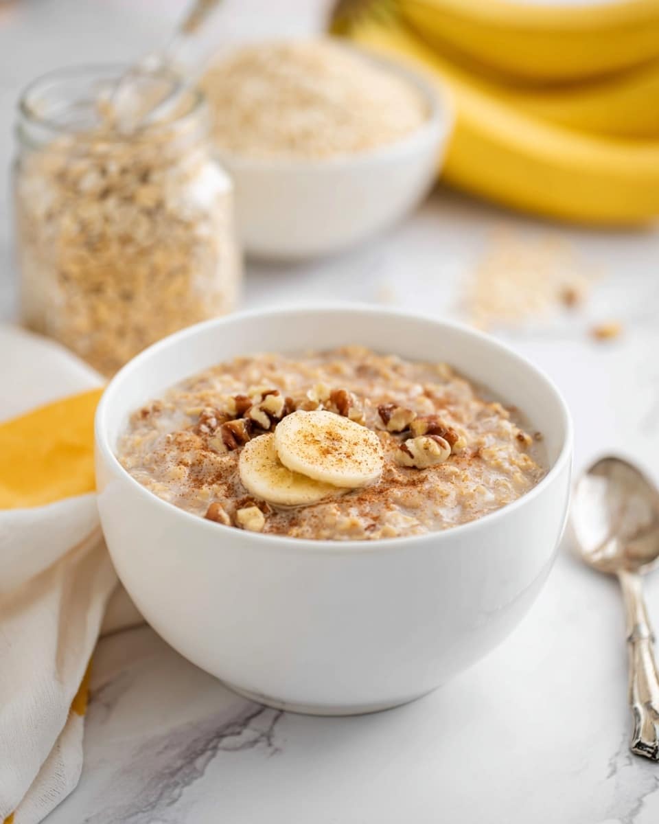 A white bowl filled with creamy oatmeal showing a soft, beige base texture mixed with small oat pieces throughout. On top are scattered dark brown pecan halves and a single light yellow banana slice dusted lightly with cinnamon. A silver spoon rests inside the bowl on the right side. The bowl sits on a white marbled surface with loose oat flakes nearby, a blurred bunch of yellow bananas in the background, and a jar with a beige smoothie partially visible to the right. photo taken with an iphone --ar 4:5 --v 7