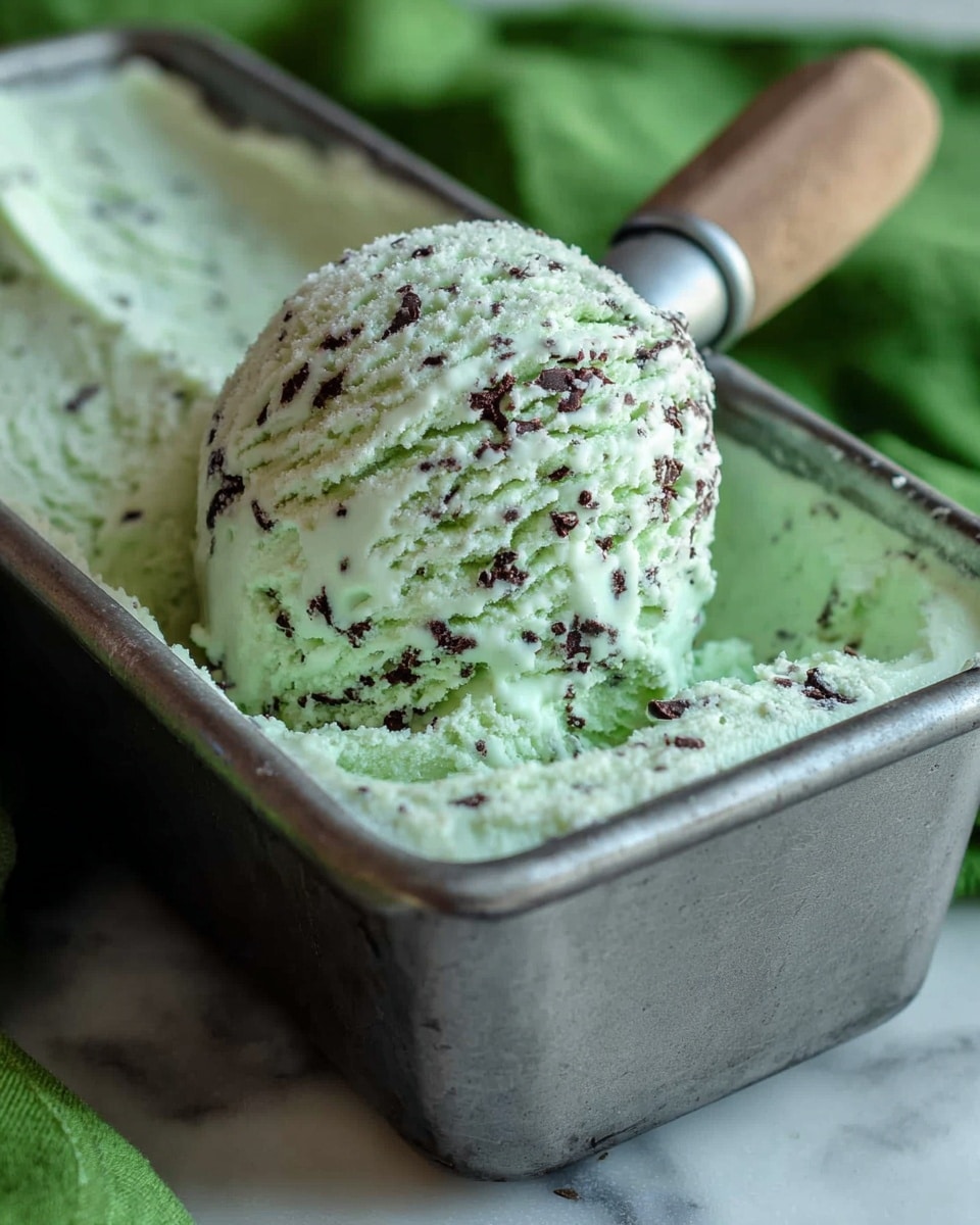A close-up image shows a single scoop of mint chocolate chip ice cream with pale green color and small dark chocolate bits inside, lifted by a metal ice cream scoop from a metal container filled with the same ice cream. The background has a soft green cloth, while the container rests on a white marbled texture surface. The ice cream texture looks creamy and slightly crumbly at the edges. Photo taken with an iphone --ar 4:5 --v 7
