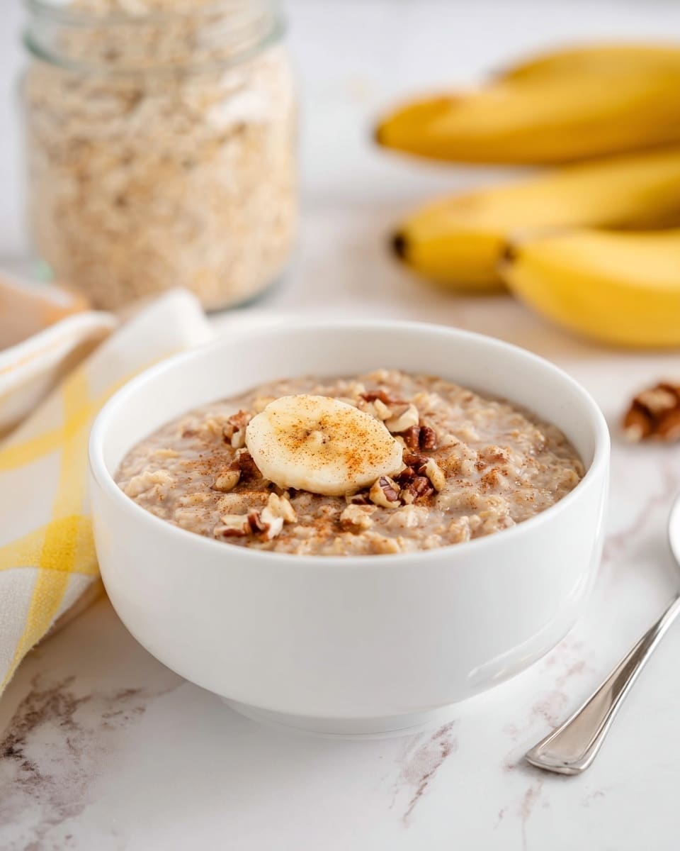 A white bowl filled with oatmeal that has a creamy texture and light brown color, topped with small pieces of nuts and a single banana slice sprinkled with cinnamon. The oatmeal appears thick and hearty. In the background, there is a blurred white bowl filled with oats, a glass jar with a metal spoon inside, and a bunch of yellow bananas. The scene is set on a white marbled surface with a white and yellow cloth nearby. Photo taken with an iphone --ar 4:5 --v 7