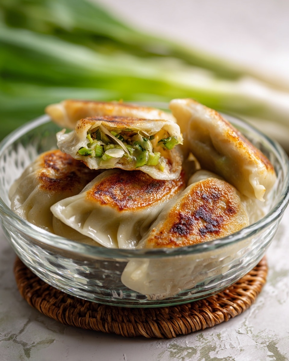 A clear glass bowl holds three half-moon shaped dumplings with a light golden-brown pan-fried crust on top. The dumpling wrappers are thin and soft, showing a slightly translucent texture. Inside, bright green vegetable filling is visible, with glossy, fresh-looking green strips peeking out from the dumpling edges. The bowl rests on a woven natural-colored coaster, with blurred green stalks in the background over a white marbled surface. photo taken with an iphone --ar 4:5 --v 7
