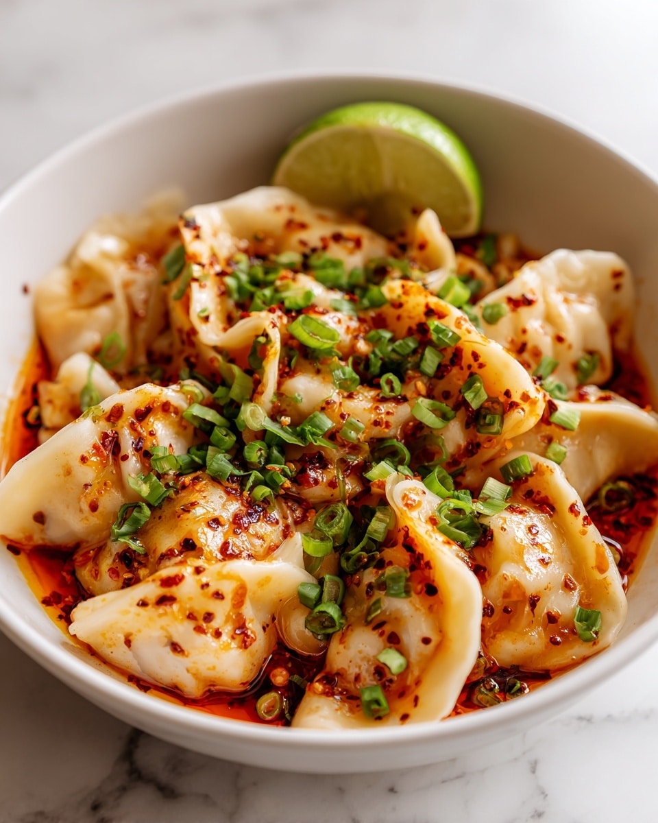 The image shows a white bowl filled with several creamy-colored dumplings covered in a light orange sauce. The dumplings have a soft and slightly wrinkled texture, topped with green herbs and small red chili flakes scattered over them. A bright green lime wedge rests on the edge of the bowl, adding a fresh color contrast. The sauce looks thick and shiny, pooling around the dumplings with some oil visible on the surface, giving it a rich appearance. The photo taken with an iphone --ar 4:5 --v 7