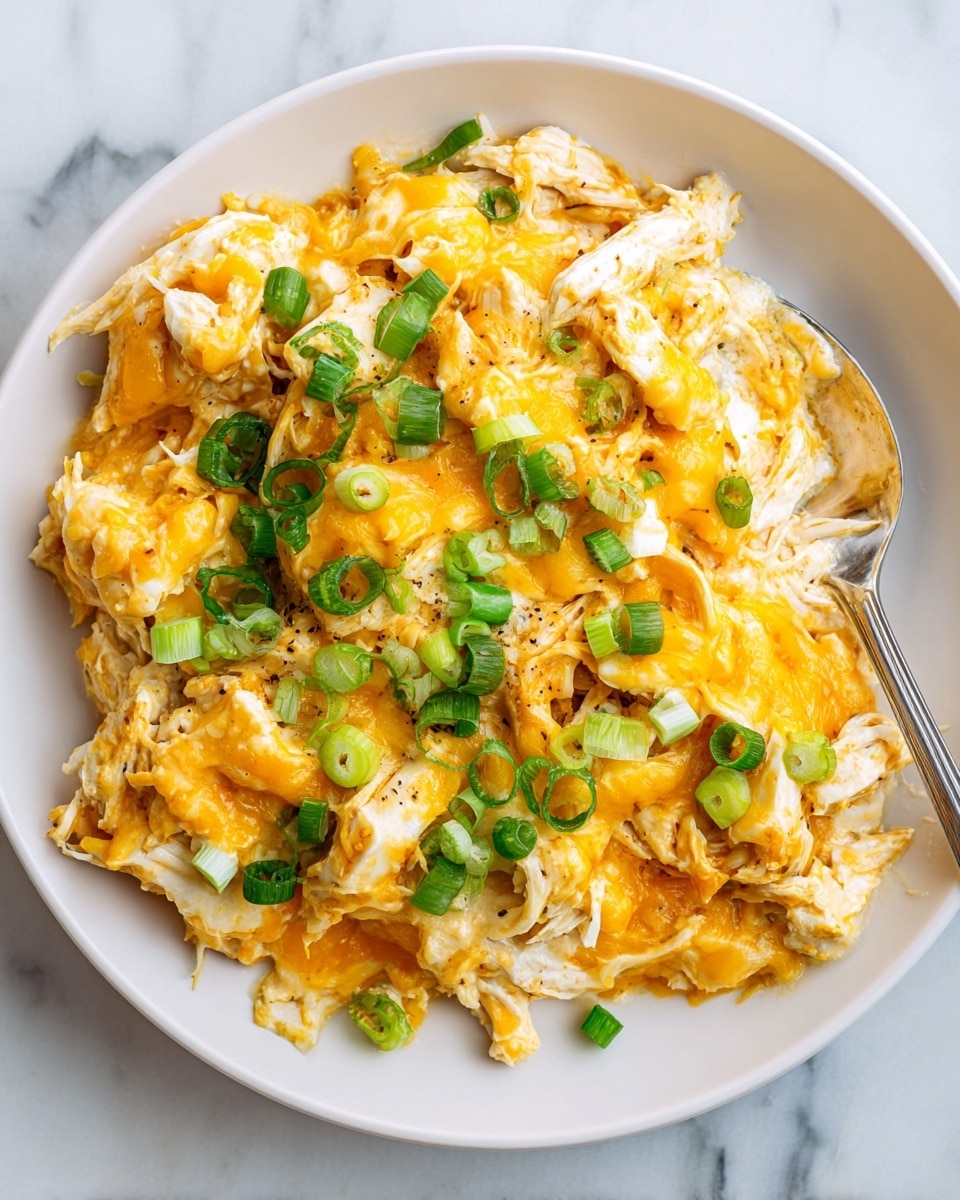 The image shows a close-up of a creamy scrambled egg dish on a white plate, layered with melted bright orange cheddar cheese evenly spread over the soft, pale yellow eggs. Scattered on top are small pieces of shredded light brown chicken mixed within the eggs. Freshly chopped green onions are sprinkled generously over the cheese, adding a vibrant green color and fresh texture contrast. The background is a white marbled texture that makes the dish stand out clearly. Photo taken with an iphone --ar 4:5 --v 7