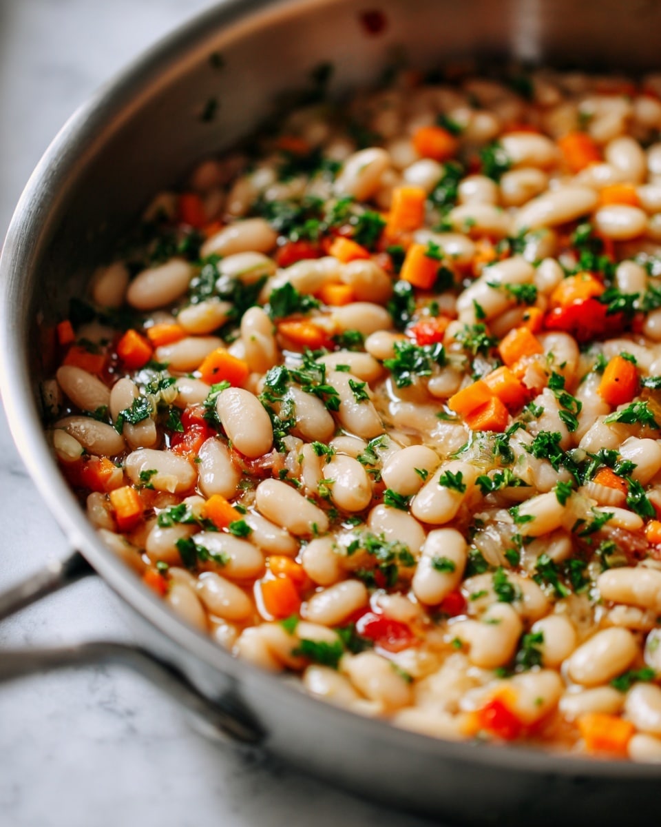 A close-up view of a skillet filled with cooked white beans mixed with small diced orange carrots, finely chopped green herbs sprinkled on top, and tiny bits of red tomato adding color. The beans are soft and creamy, scattered evenly across the pan with light steam rising. The mixture shows a variety of textures from the smooth beans, crunchy vegetables, and fresh herbs, all blending naturally together. The skillet sits on a white marbled surface. photo taken with an iphone --ar 4:5 --v 7