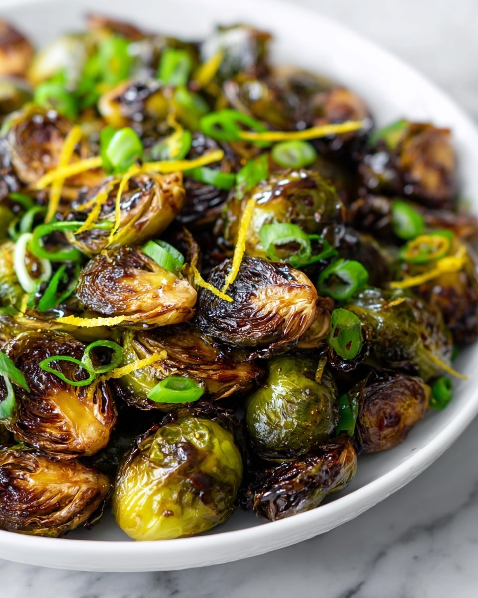 The image shows a close-up of roasted Brussels sprouts that are caramelized and browned with a glossy texture from a honey balsamic glaze. The sprouts are halved and appear layered, with dark, crispy edges and vibrant green inner leaves. Scattered on top are thin slices of fresh green onions, adding a pop of bright green color. There are also small bits of light yellow lemon zest sprinkled over the dish, giving a fresh contrast to the darker tones of the Brussels sprouts. The dish sits on a white plate over a white marbled surface. photo taken with an iphone --ar 4:5 --v 7