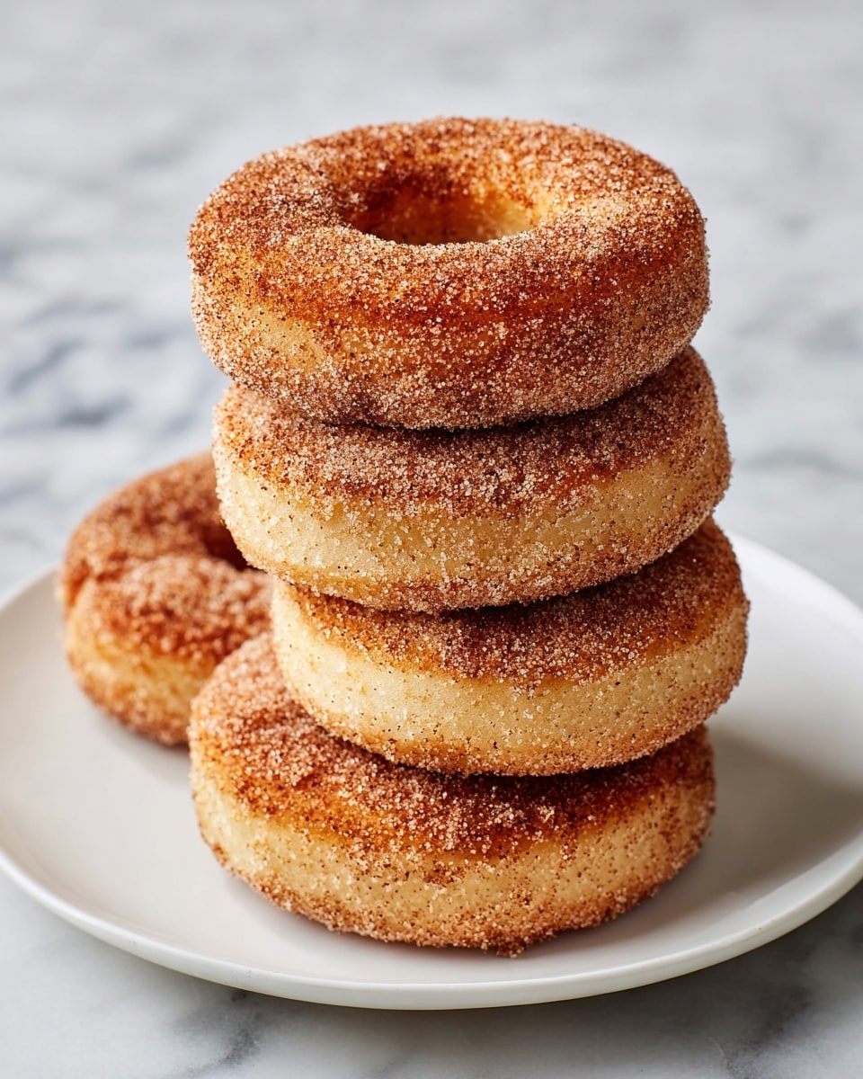 A white plate holds five cinnamon sugar-coated doughnuts stacked neatly, each doughnut showing a golden-brown crispy texture with a rough sugary surface. The cinnamon sugar mix appears as a light brown granular layer covering the entire outer ring of each doughnut. The dough itself is soft and slightly porous, with the edges showing a slight darker crispiness. The plate rests on a white marbled surface. photo taken with an iphone --ar 4:5 --v 7