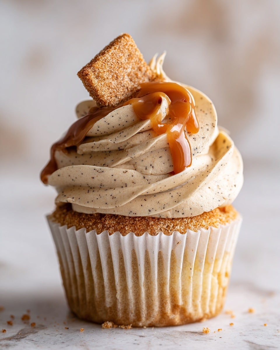 The image shows a close-up of a cupcake placed on a white paper liner, sitting on a white marbled surface. The cupcake has a light brown base topped with three swirls of thick, creamy frosting with a speckled texture, likely cinnamon or spice flavored. Over the frosting, there is a thin layer of golden caramel sauce that drips slightly down the sides. On top of the caramel, a small, square piece of cinnamon sugar-coated fried dough or pastry is placed, adding a crunchy texture. The background features more cupcakes, softly blurred to keep focus on the front cupcake. photo taken with an iphone --ar 4:5 --v 7