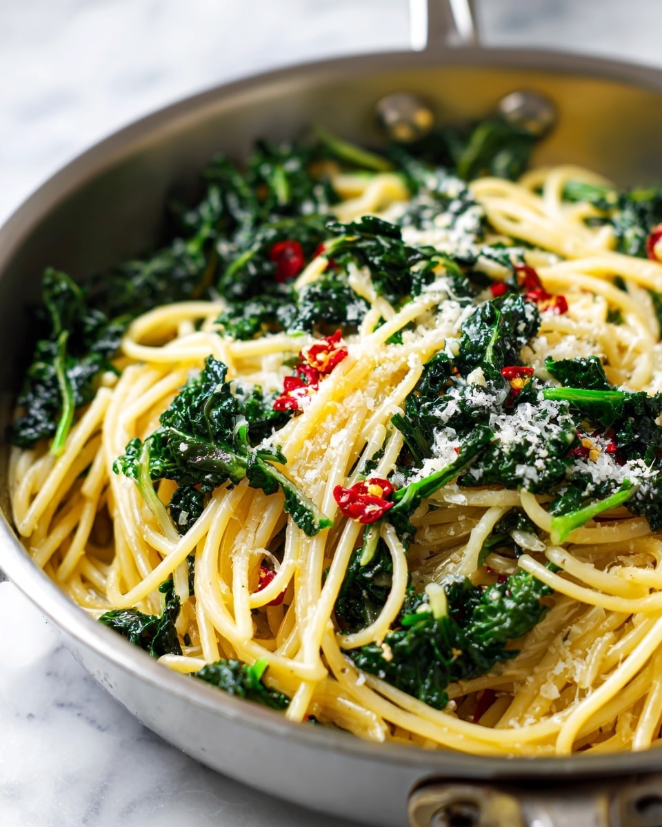 A close-up view of a pasta dish in a silver pan placed on a white marbled surface. The dish has a base layer of light yellow spaghetti noodles, topped with bright dark green pieces of cooked kale spread evenly throughout. On top of the kale and noodles, there are light sprinkles of white grated cheese and small red chili flakes scattered across for color contrast. The textures show the smoothness of the spaghetti and the roughness of the kale leaves. Photo taken with an iphone --ar 4:5 --v 7