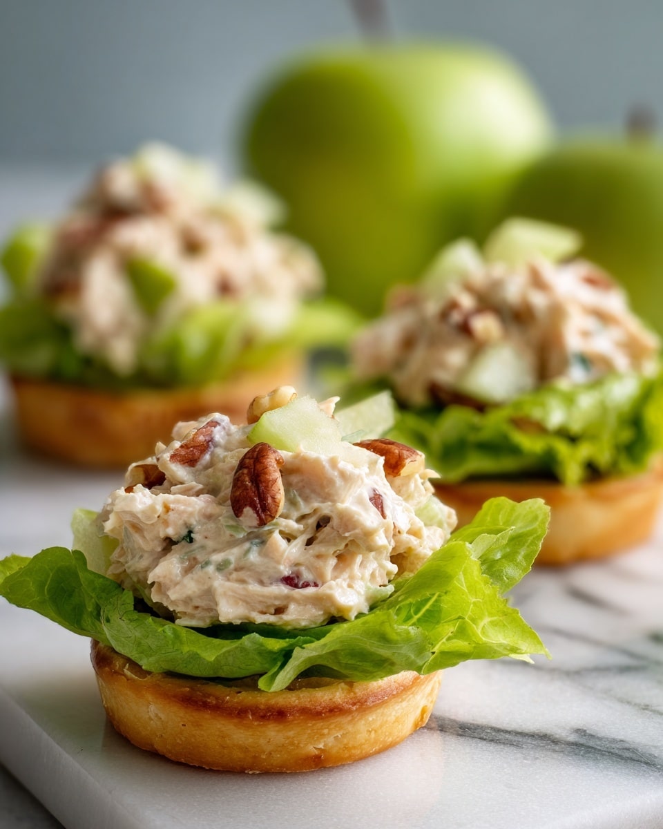 The image shows three round tartlets placed on a white marble surface. Each tartlet has a golden-brown crispy base at the bottom, topped with fresh bright green leafy lettuce. On top of the lettuce, there is a generous scoop of creamy chicken salad mixture, which is white with visible small colorful bits of red apples, light green celery pieces, and brown nuts. The texture of the salad looks soft and chunky. In the background, a fresh green apple is visible out of focus. Photo taken with an iphone --ar 4:5 --v 7