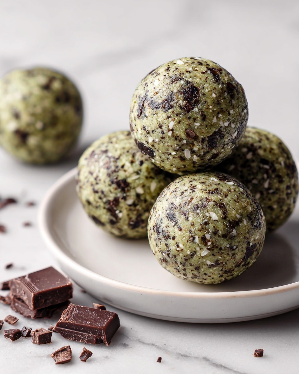 The image shows a close-up of five round energy balls with a speckled texture, placed on a white plate on a white marbled surface. Each ball has a smooth greenish base with bits of dark brown chocolate pieces and small white flakes mixed throughout. The balls are stacked with one in the front, in sharp focus, and four more slightly blurred in the background. Around the plate, there are several small chunks of broken dark chocolate scattered casually. photo taken with an iphone --ar 4:5 --v 7