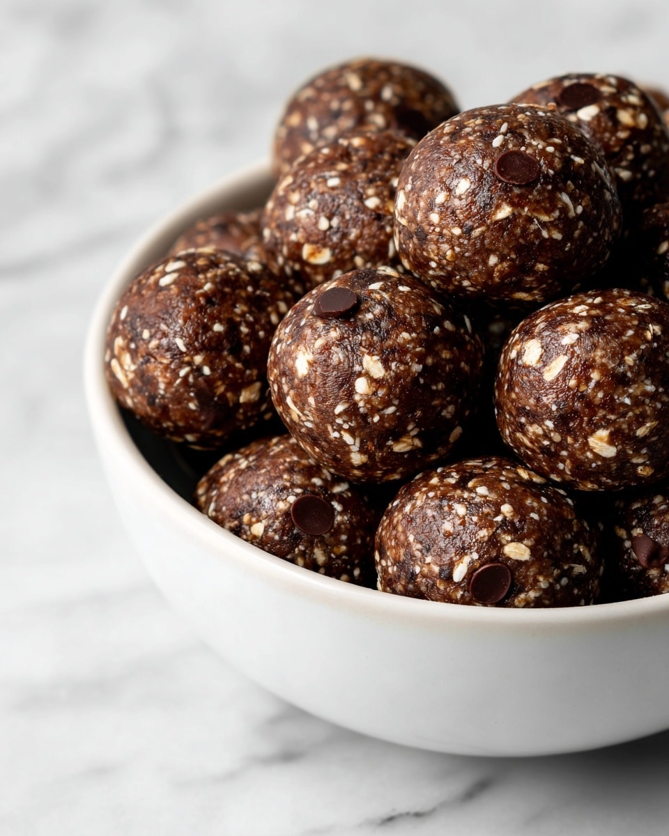 A close-up view of many small round balls made of dark brown, sticky oat mixture with visible oatmeal flakes and small dark chocolate chips mixed throughout. The balls are stacked together inside a white bowl that fills the frame, resting on a white marbled surface. The texture looks soft and chewy, with a slightly glossy finish on each ball. photo taken with an iphone --ar 4:5 --v 7