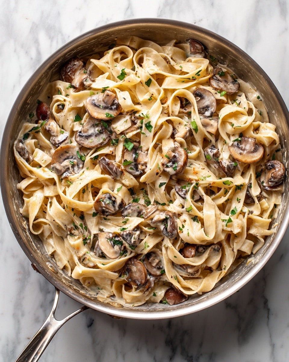 The image shows a close-up of a creamy pasta dish in a white pan sitting on a white marbled surface. The dish has flat, wide noodles coated in a light beige cream sauce, mixed with sliced mushrooms that are dark brown and gray. Small green herb pieces are scattered throughout the sauce, adding a touch of color. The sauce looks smooth and slightly thick, clinging to the noodles and mushrooms evenly. Photo taken with an iphone --ar 4:5 --v 7