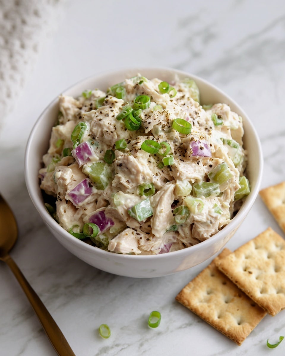 A white bowl filled with creamy chicken salad sits on a white marbled surface. The salad has shredded white chicken mixed with chopped green celery and bits of purple onion, all coated in a light, creamy dressing. The top is sprinkled with chopped green onions and a dash of pepper for extra texture and color. Around the bowl, there are several square saltine crackers. A beige linen cloth is partially visible on the right side. photo taken with an iphone --ar 4:5 --v 7